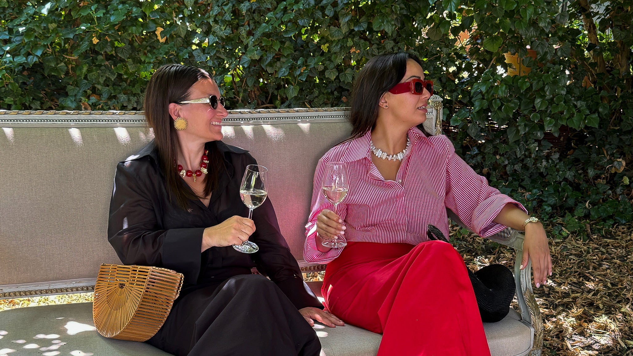 Zwei Frauen in eleganter Kleidung aus der Kollektion The New Luxury von PALAIS du SOLEIL – Kampagnenbild mit Sektgläsern auf einem Sofa im Garten