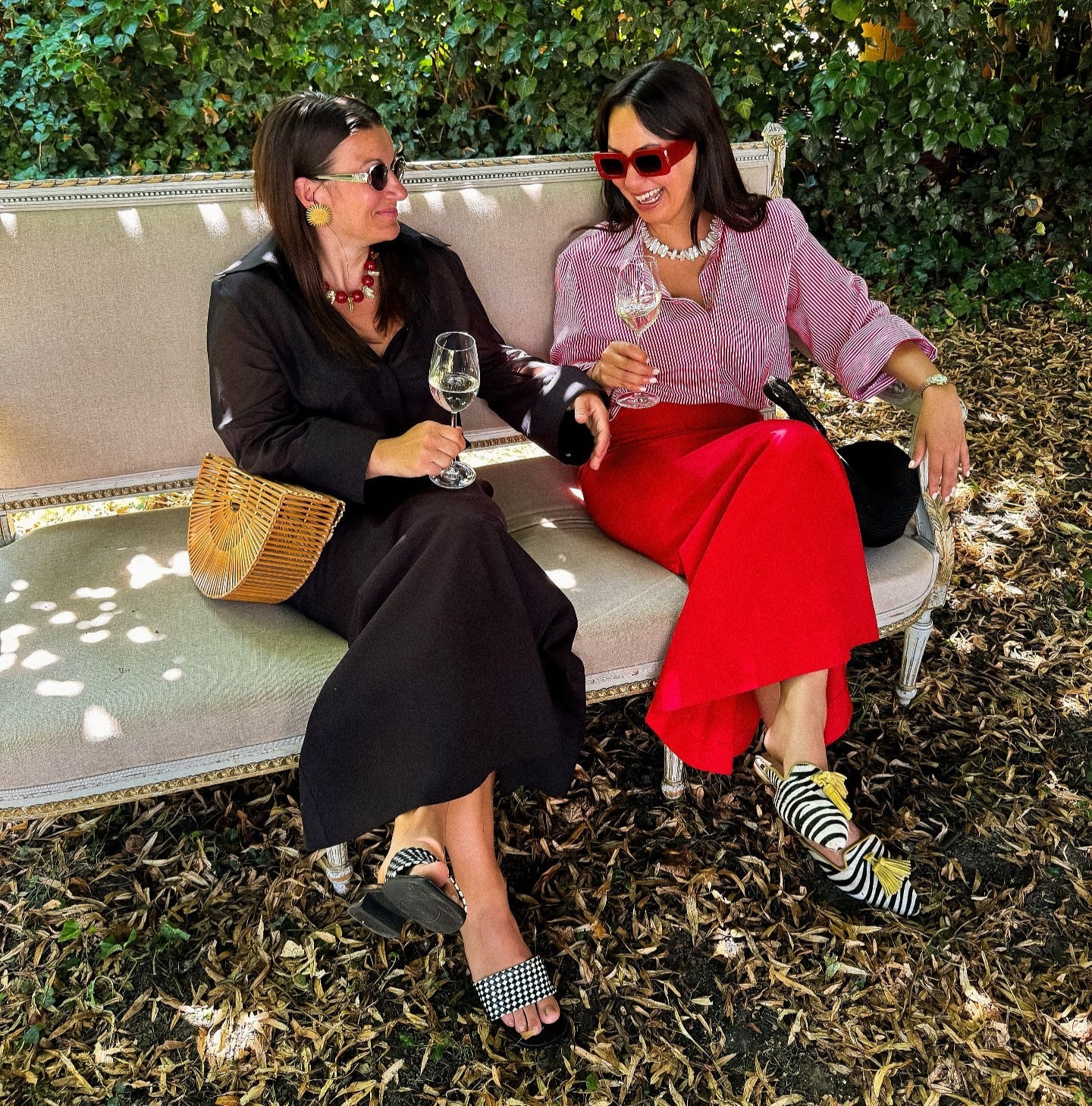 Zwei Frauen in eleganter Kleidung aus der Kollektion The New Luxury von PALAIS du SOLEIL – Kampagnenbild mit Sektgläsern auf einem Sofa im Garten